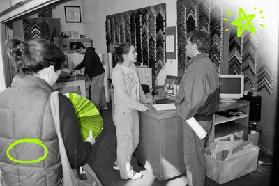 Wide black-and-white snapshot of a busy frame shop interior in 2001, shot from the doorway. Four people at different depths — a waiting customer near the camera leafing through a fan of moulding samples tinted acid green, a staff member mid-conversation with a customer at the counter, and an employee bent over the mat cutter at the back wall.