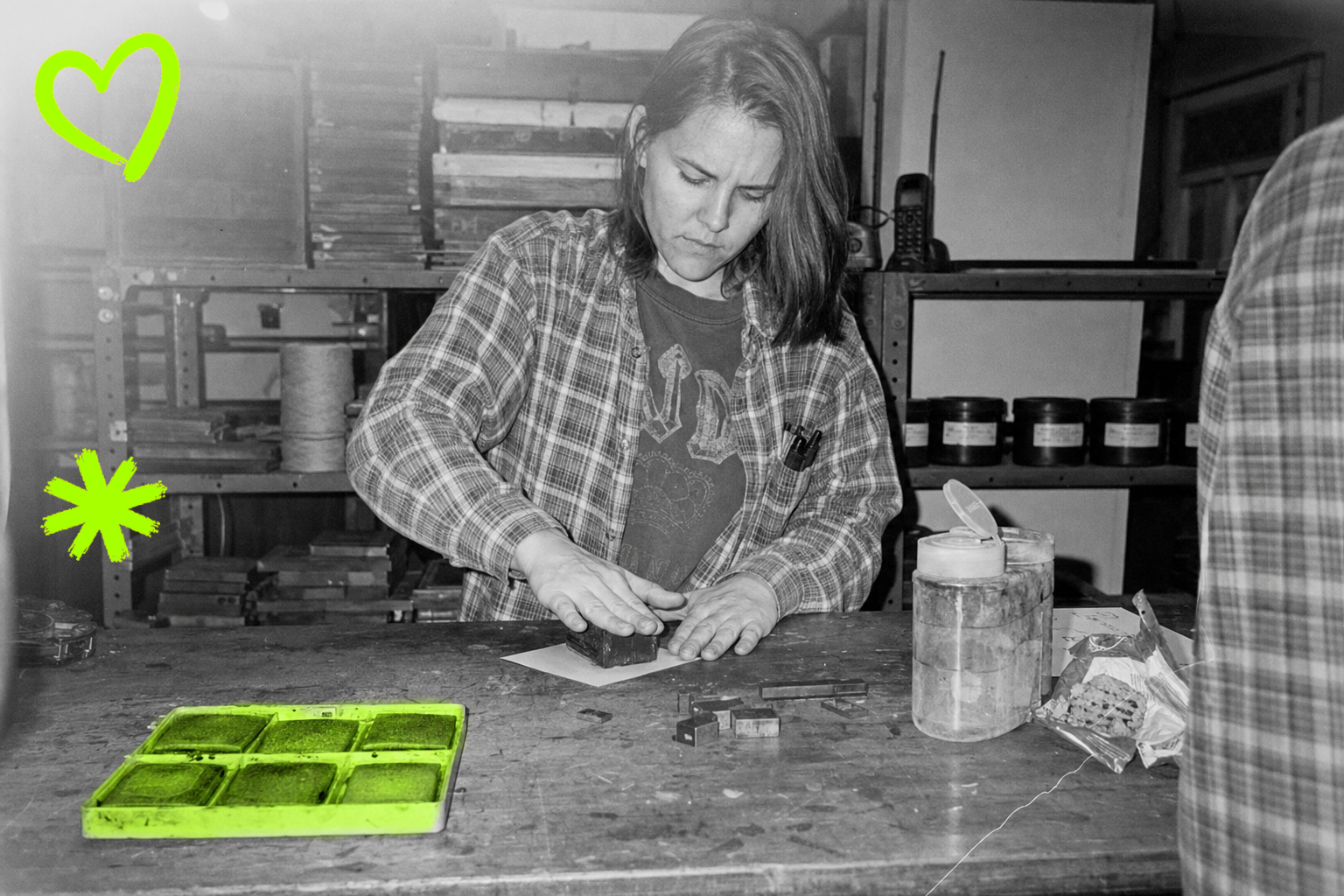 Black-and-white point-and-shoot photograph from 1998: a young woman in a flannel shirt over a faded tee presses a rubber stamp block against a proof sheet at a cluttered letterpress bench, a segmented ink-pad tray in the foreground highlighted in acid green