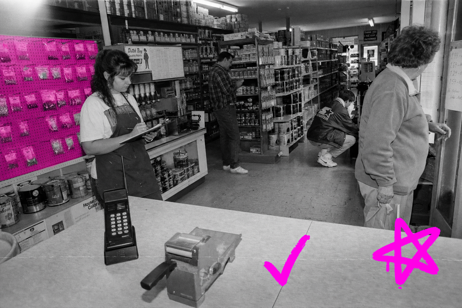 Wide black-and-white interior of a small 1993 hardware store: four people at different depths go about their work among shelves of paint cans and fasteners, a manual credit-card imprinter on the counter, a hot-magenta-tinted pegboard of bolt bags on the left wall.