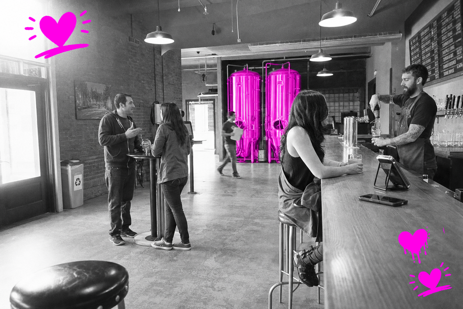 Wide black-and-white point-and-shoot photograph inside a craft brewery taproom in 2014: a tattooed bartender pours a pint at a wooden bar while a woman on a barstool holds her glass and two customers talk at a high-top table in the middle ground; a blurred figure moves near two large fermentation tanks in the background, which glow with a translucent hot-magenta wash.