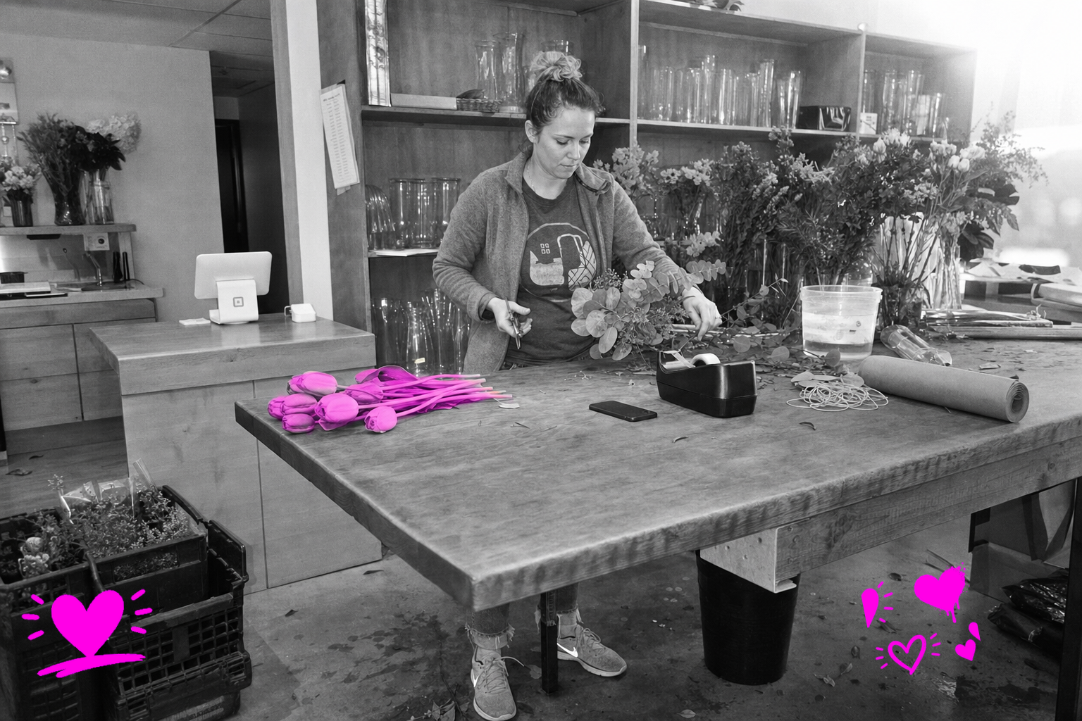 Black-and-white point-and-shoot photo of a young woman mid-arrangement at a florist's worktable, surrounded by cut stems, a kraft paper roll, a card reader on a tablet, and glass vases on shelving behind her — a loose bunch of tulips on the table washed in translucent hot magenta.